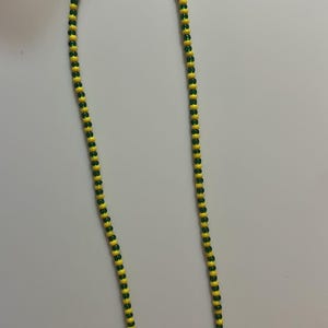 May include: A beaded necklace featuring alternating yellow and green beads. The beads are small and strung together to form a long, flexible strand. The necklace is displayed against a plain, light-colored background.