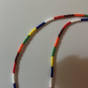 May include: A colorful beaded necklace with sections of white, red, brown, orange, green, yellow, and blue beads. The necklace is displayed against a plain white background. The beads are small and round.