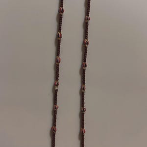 May include: Two strands of beaded jewelry. The strands are dark brown with alternating decorative beads featuring red, white, and black stripes. The smaller beads are also dark brown. The jewelry hangs against a plain, off-white background.