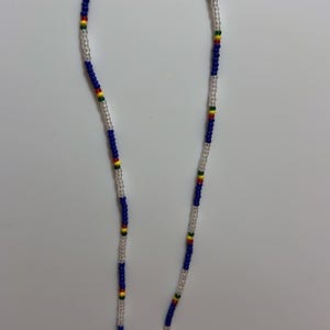 May include: A beaded necklace featuring a pattern of blue, clear, red, yellow, and green beads. The necklace is made of small, round beads strung together. The beads are arranged in a repeating pattern of blue and clear beads with accents of red, yellow, and green.
