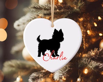 Puede incluir: Adorno navideño blanco en forma de corazón con una silueta negra de perro y el nombre "Charlie" en escritura roja. Cuelga de una cuerda dorada, sobre un fondo borroso de árbol de Navidad.