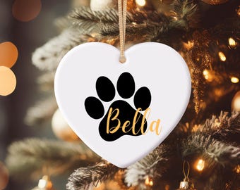 Puede incluir: Adorno navideño blanco en forma de corazón con una huella de pata negra y el nombre "Bella" en letras doradas. El adorno cuelga de una cuerda dorada sobre un fondo borroso de árbol de Navidad.