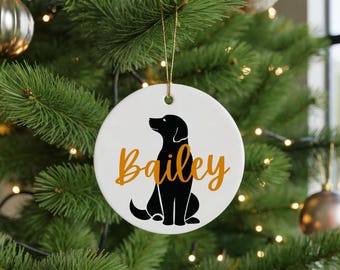 May include: White ceramic Christmas ornament with a black silhouette of a dog and the name "Bailey" in orange script. The ornament hangs from a gold string and is displayed on a Christmas tree with other ornaments.