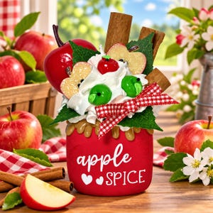 May include: A red candle jar with the text apple SPICE and two white hearts. The top features faux whipped cream, a red apple, green apple slices, cinnamon sticks, green leaves, and a red and white checkered ribbon bow.