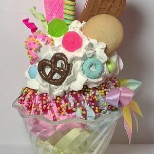 May include: A whimsical dessert sundae in a clear glass bowl, featuring layers of pink and blue ice cream, whipped cream, and colorful sprinkles. Toppings include a pretzel, cookie, and various candies, with a decorative bow.