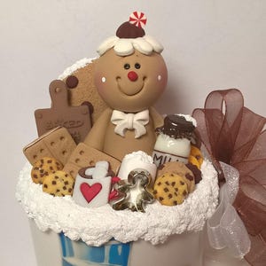 May include: A whimsical gingerbread man figurine with a white bow tie, surrounded by cookies, a milk bottle, and a mug with a heart. The figurine is topped with a peppermint candy and sits in a white cup with blue accents. A brown ribbon is tied to the side.