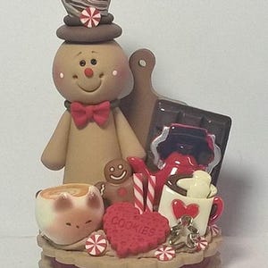 May include: A whimsical gingerbread man figurine with a top hat, bow tie, and rosy cheeks. The scene includes a mug, heart-shaped cookie with "COOKIES" text, chocolate, and candy canes. A festive holiday decoration.