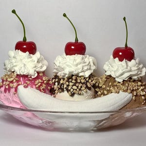 May include: A classic banana split dessert with three scoops of ice cream: pink, white, and brown. Each scoop is topped with whipped cream, nuts, and a maraschino cherry. The split sits in a clear glass boat.