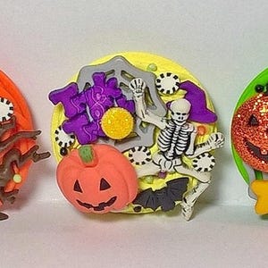 May include: Three Halloween-themed decorative buttons. The first features a ghost, spiderweb, and the words "Trick or Treat." The second has a pumpkin, skeleton, and bat. The third includes a jack-o'-lantern, spider, and witch. Each button is round with vibrant colors.