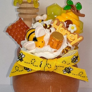 May include: A round wooden pot decorated with a yellow ribbon featuring bee patterns. The top is adorned with white whipped cream, a wooden honey dipper, a honeycomb piece, a small bee, a honey jar, and a sign with the word Honey.