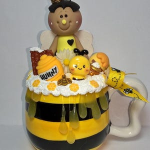 May include: A whimsical, hand-painted ceramic mug with a bee theme. The mug features yellow and black stripes, with a decorative bee figurine on top. The mug is adorned with honey drips and the word "HUNNY".