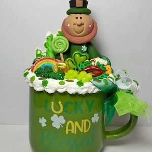 May include: Green ceramic mug with a leprechaun figurine on top. The mug has white frosting around the rim and is decorated with green shamrocks, gold coins, and a rainbow. The mug says "Lucky and Blessed" in yellow lettering.