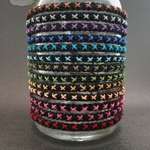 May include: A stack of colorful bracelets with black stitching, displayed in a glass jar. The bracelets are made with a variety of colors, including red, orange, yellow, green, blue, purple, and white.