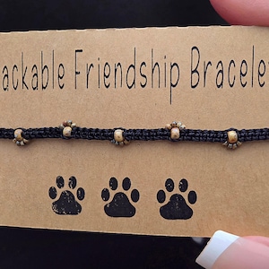May include: A black macrame friendship bracelet with small, decorative beads. The bracelet is displayed on a brown card with the text "Stackable Friendship Bracelet" and paw print graphics.
