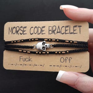 May include: A black and gold bracelet with a skull charm. The bracelet is made of black cord and gold beads. The bracelet is labeled "Morse Code Bracelet" and the words "Fuck" and "Off" are spelled out in Morse code.