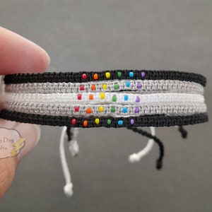 May include: A multi-colored friendship bracelet with a rainbow pattern of beads. The bracelet is made of black, white, and gray string.
