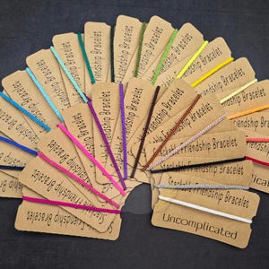 May include: A collection of thin, colorful cord bracelets arranged in a fan shape. Each bracelet is attached to a rectangular brown card printed with the text Stackable Friendship Bracelet and Uncomplicated in black ink.