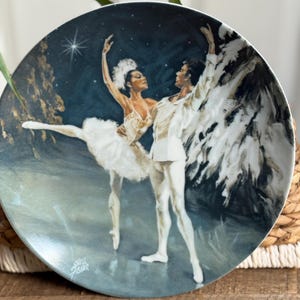 May include: Decorative plate featuring a ballet scene with a male and female dancer in white costumes. The background is a dark blue with a star and a tree. The plate has a gold rim and a signature in the bottom left corner.