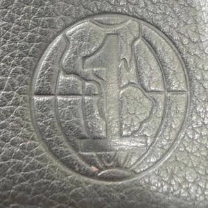 May include: Close-up of a black leather surface with a circular embossed design. The design features a large number "1" in the center, surrounded by a globe-like pattern within a circular border. The leather has a textured appearance.