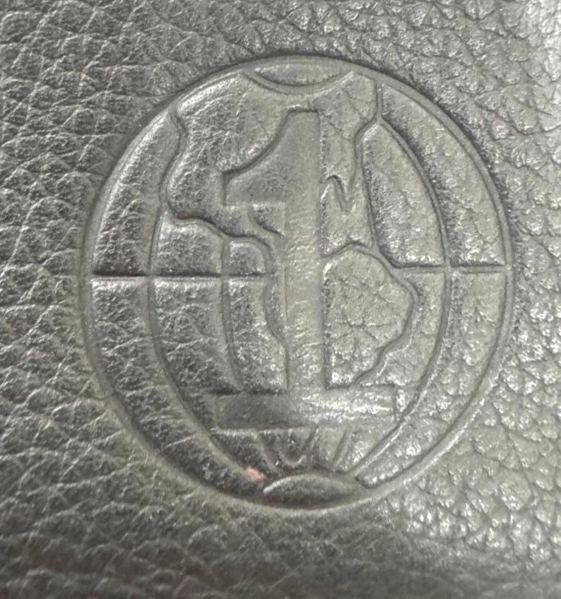 May include: Close-up of a black leather surface with a circular embossed design. The design features a large number "1" in the center, surrounded by a globe-like pattern within a circular border. The leather has a textured appearance.