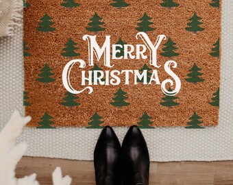 May include: A rectangular doormat with a brown background and a pattern of green Christmas trees. The words "MERRY CHRISTMAS" are printed in white, with decorative flourishes. Black boots and blue jeans are visible at the bottom.