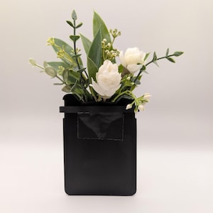 May include: A black rectangular vase holding white flowers and green foliage. The arrangement features white blooms, green leaves, and sprigs of greenery, creating a natural and elegant display. The vase has a simple, modern design.