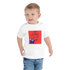 May include: White toddler t-shirt with a red square graphic. The graphic features a cartoon police officer and the text "Stop Bullying it ain't right" in black script. The child is wearing blue jeans.