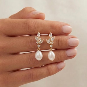 May include: Gold drop earrings with a leaf design, clear crystals, and a teardrop pearl. The earrings are held up by a hand, highlighting the intricate details and elegant style of the jewellery.
