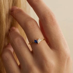 May include: A gold ring with two pear-cut gemstones, one sapphire blue and one clear, set together. The ring is on a finger, highlighting the gemstones and the slender gold band. The ring's design is simple and elegant.