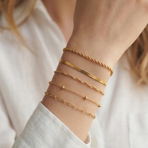 May include: A collection of gold bracelets in various styles, including a rope chain, a flat snake chain, a delicate chain with small beads, and a paperclip chain. The bracelets are worn on a wrist against a white shirt.