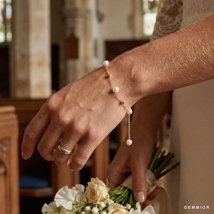 May include: A delicate gold bracelet with pearls and small gold beads. The bracelet is worn on a wrist, with a wedding ring visible on the finger. A bouquet of white and cream flowers is held in the hand.