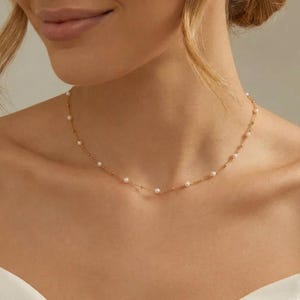 May include: A fine gold necklace adorned with small, evenly spaced pearls. The necklace is worn around a person's neck, highlighting its elegant design. The pearls are white and the chain is gold.