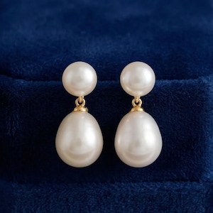 Può includere: Un paio di orecchini pendenti con perle. Ogni orecchino presenta una piccola perla rotonda nella parte superiore, collegata da un elemento dorato a una perla più grande a forma di goccia nella parte inferiore. Le perle sono di un bianco crema lucido.
