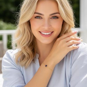 Op de afbeelding: Een delicate gouden armband met een kleine, donkerblauwe edelsteen. De armband heeft een dunne gouden ketting. De edelsteen is rond en centraal geplaatst. De persoon draagt een lichtblauw overhemd.