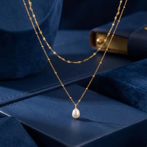May include: A layered gold necklace with a small, white pearl pendant. The necklace has two strands, one with small beads and the other with a chain design. The jewelry is displayed on a dark blue surface.