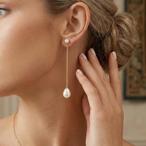 May include: A close-up of a gold chain earring with a white pearl at the end. The earring is dangling from the ear. The earring is a drop style. The background is blurred.