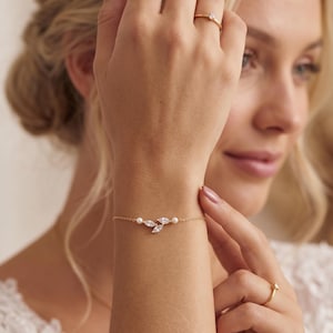 Könnte beinhalten: Ein zartes goldenes Armband mit einem Blattdesign aus klaren Kristallen und kleinen Perlen. Das Armband wird am Handgelenk getragen, und ein goldener Ring mit einem kleinen Diamanten befindet sich am Finger. Der Schmuck ist elegant und für besondere Anlässe geeignet.
