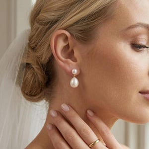 May include: Elegant pearl drop earrings. The earrings feature a small, round pearl at the top and a larger, teardrop-shaped pearl suspended below. The setting appears to be gold. The image shows the earrings on a person's ear, with a blurred background.