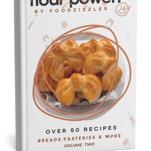 May include: A cookbook titled "flour power." features a basket of golden-brown baked goods. The cover includes the text "OVER 50 RECIPES BREADS, PASTRIES & MORE VOLUME TWO" and the author's name, "BY FOODZIZZLES."