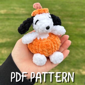 May include: A small, white, plush crochet dog figure with black ears and a black nose, wearing a textured orange pumpkin-shaped body and a matching orange hat with a small stem on top. The text PDF PATTERN is printed at the bottom of the image.