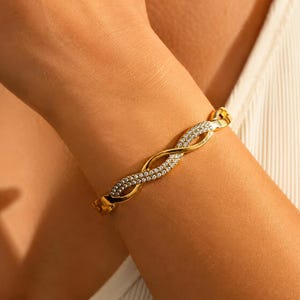 May include: A gold-coloured bracelet featuring a woven pattern, embellished with clear crystals. The bracelet is fastened and worn on a wrist. The jewellery item has a polished finish, reflecting light.