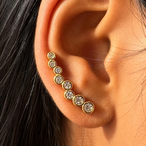 May include: A gold ear climber earring featuring seven round, bezel-set clear stones arranged in a curved, ascending line that follows the natural shape of the ear cartilage. The metal has a polished yellow gold finish.