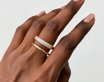 May include: A gold ring set consisting of two separate bands. The top band features a pavé setting with numerous small, clear, sparkling stones, while the bottom band is a smooth, plain gold ring with a rounded, polished finish.