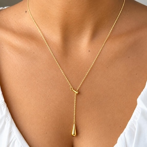 May include: A gold-toned lariat necklace featuring a delicate cable chain and a single smooth, teardrop-shaped pendant suspended from a small cylindrical connector. The necklace has a polished metallic finish and a Y-shaped drop design.
