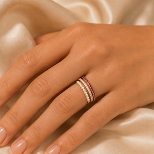 May include: A stack of three thin rings worn on a finger. The first ring features a row of small white pearls, the second ring is set with clear sparkling stones, and the third ring is set with a continuous row of small, vibrant red stones.