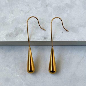 May include: A pair of gold-toned drop earrings featuring a smooth, polished teardrop-shaped pendant suspended from a curved wire hook. The earrings have a reflective metallic finish and a minimalist design, showcasing a simple, elongated silhouette.