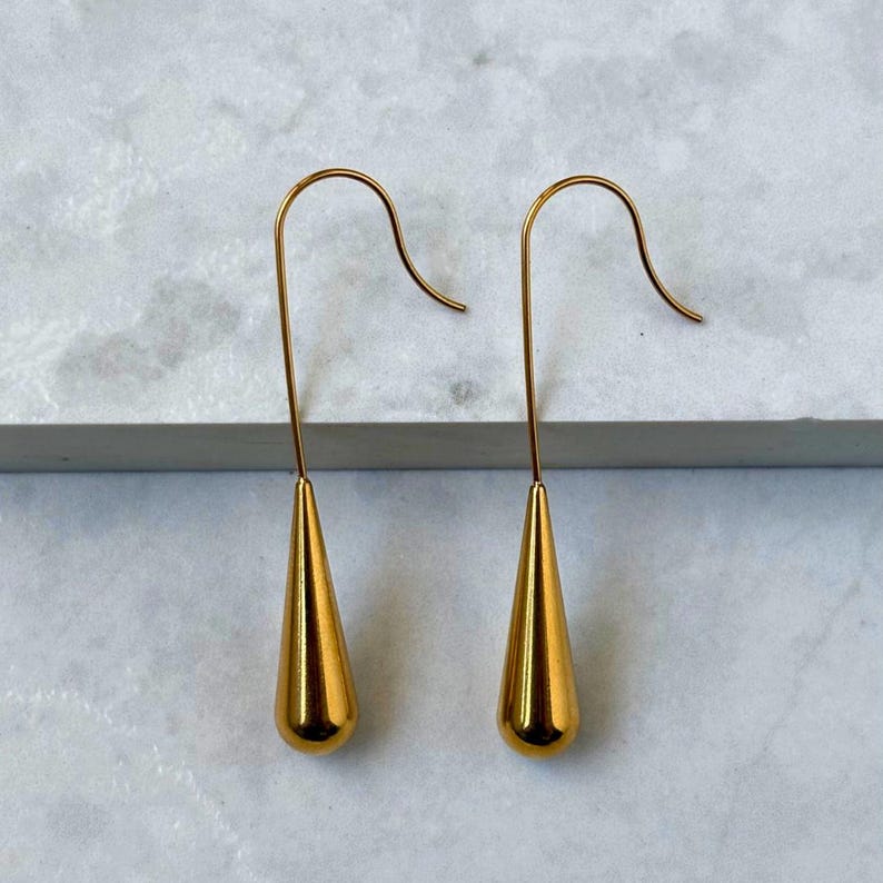 May include: A pair of gold-toned drop earrings featuring a smooth, polished teardrop-shaped pendant suspended from a curved wire hook. The earrings have a reflective metallic finish and a minimalist design, showcasing a simple, elongated silhouette.