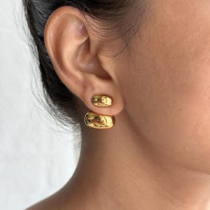 May include: A pair of gold-colored, rounded stud earrings. The earrings have a smooth, polished surface and a simple, elegant design. The earrings are shown on an ear, highlighting their size and shape.