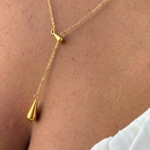 Pode incluir: Um colar dourado com um pingente em forma de gota. O colar tem uma corrente delicada e um design em forma de Y. O pingente tem um formato de gota liso e sólido. O colar é usado em um fundo de tom de pele.