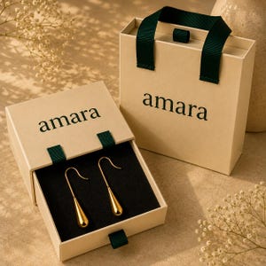 May include: A pair of gold-toned teardrop earrings with hook backings, presented in a beige sliding drawer box with a dark green ribbon pull. The box features the word amara printed in black. A matching beige gift bag with dark green handles is included.